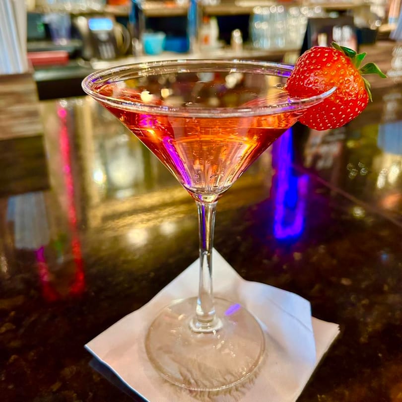 A vibrant pink cocktail with a fresh strawberry garnish sits on a bar at White Wolf Cafe.