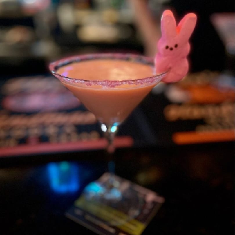 A martini glass with a pink-rimmed, orange drink and a pink Peeps bunny garnish.