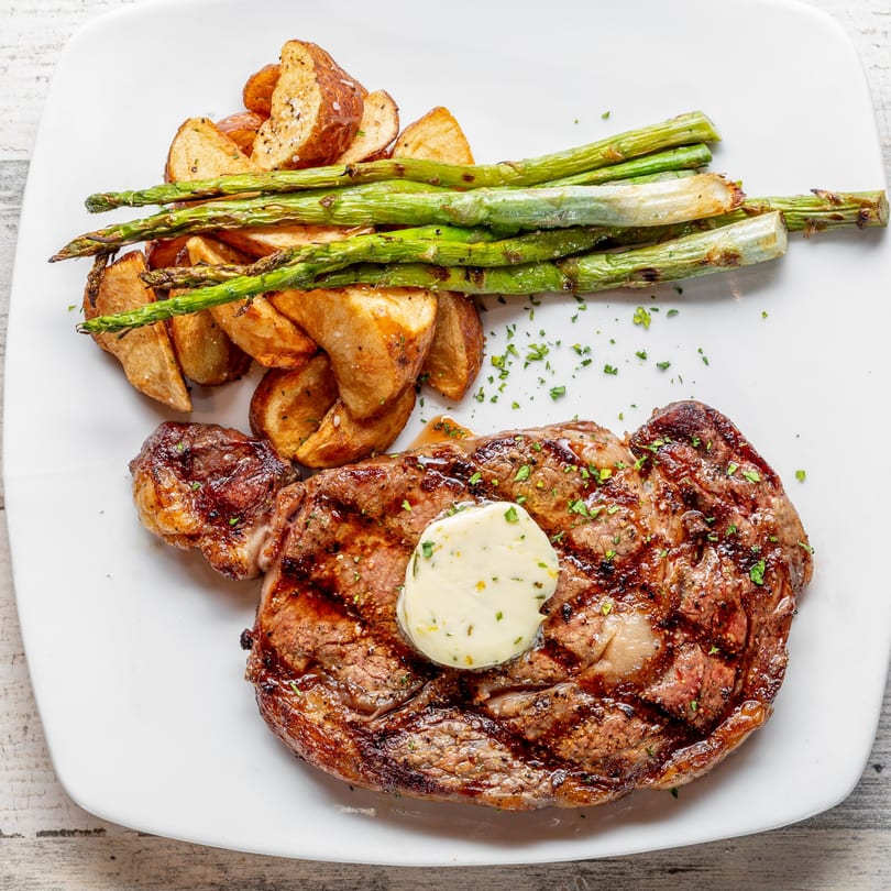 Grilled steak with herb butter, roasted potato wedges, and asparagus on a white plate.