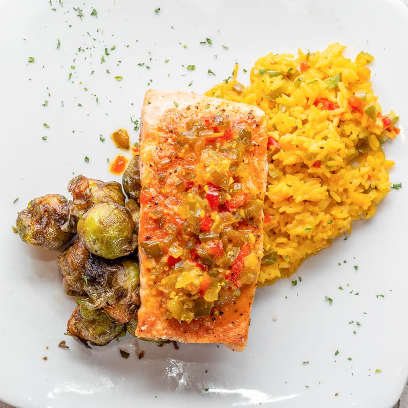Salmon fillet with colorful salsa, yellow rice, and roasted Brussels sprouts on a white plate.
