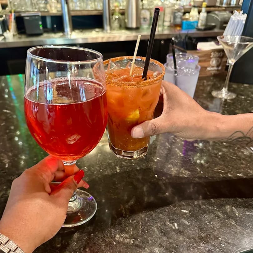 Two hands clinking glasses, one with red wine, the other with a Bloody Mary, at a bar.